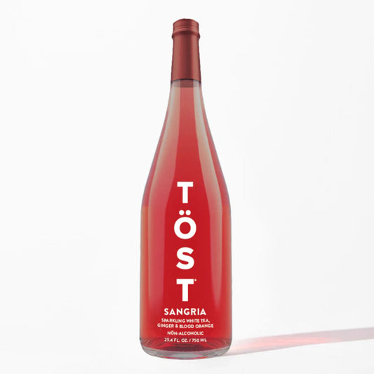 A sleek, clear glass bottle of TÖST Sangria, a non-alcoholic sparkling white tea with ginger and blood orange, stands on a light gray surface against a bright white background. The bottle is filled with a vibrant, translucent red liquid. A minimalist white label runs vertically down the center of the bottle, displaying the brand name "TÖST" in bold, stacked lettering with two dots above the "O". Below the brand name, "SANGRIA" is printed in a smaller red sans-serif font.