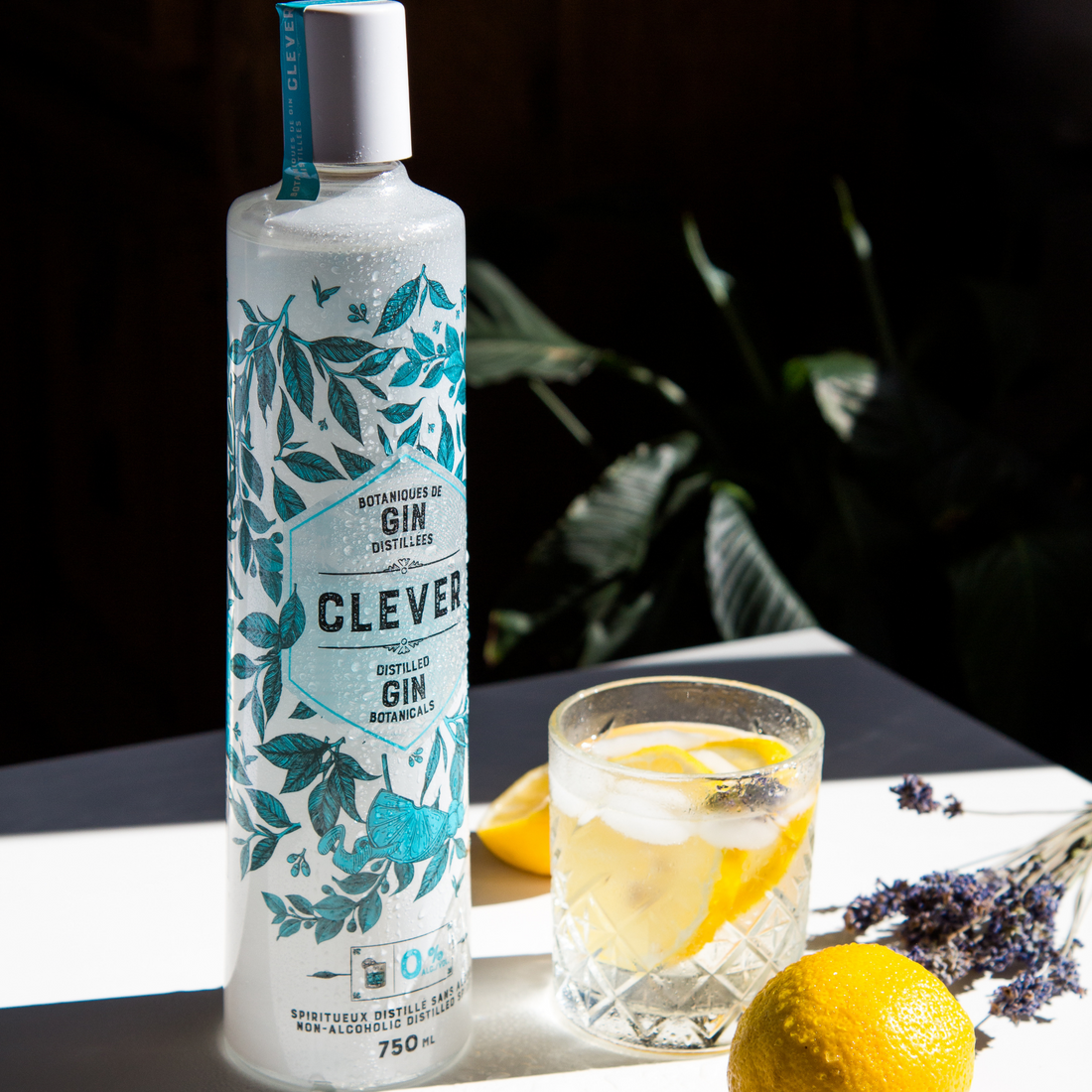 A photo of a bottle of Clever Distilled Gin Botanicals, lavender garnish, lemons, and a lavender collins drinks.