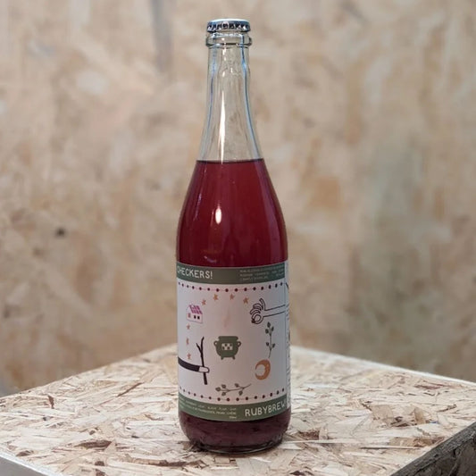A clear glass bottle of Checkers! Rubybrew non-alcoholic fermented beverage with a light brown label featuring illustrations of a house, a plant in a pot, and stars, stands on a wooden surface. The liquid inside is a deep red color.