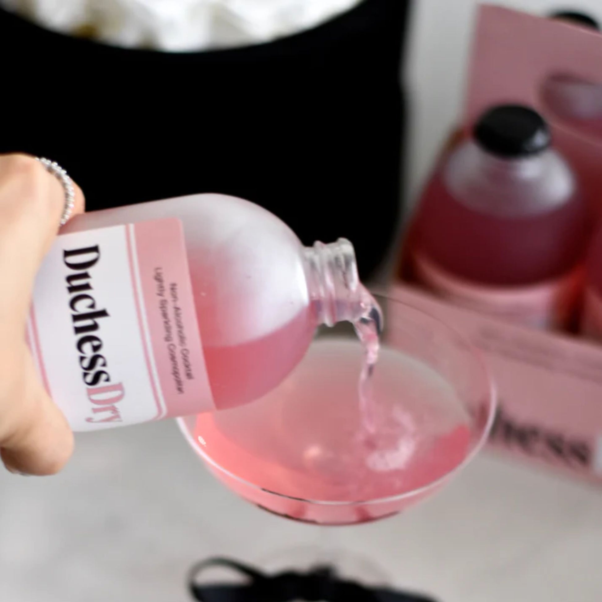 The image shows a hand pouring a pink liquid from a bottle of the Lightly Sparkling Cosmopolitan DuchessDry Non-Alcoholic Cocktail into a martini glass. The bottle is frosted glass with a pink and black label prominently displaying the DuchessDry logo. The background is blurred, suggesting a table setting.