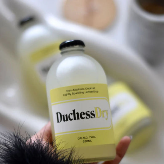 The image shows a hand holding a bottle of DuchessDry Non-Alcoholic Cocktail, specifically the Lightly Sparkling Lemon Drop flavor. The bottle is made of frosted glass and has a black cap. The label on the bottle is yellow and black, with the DuchessDry logo prominently displayed. The background is blurred, suggesting a kitchen setting.