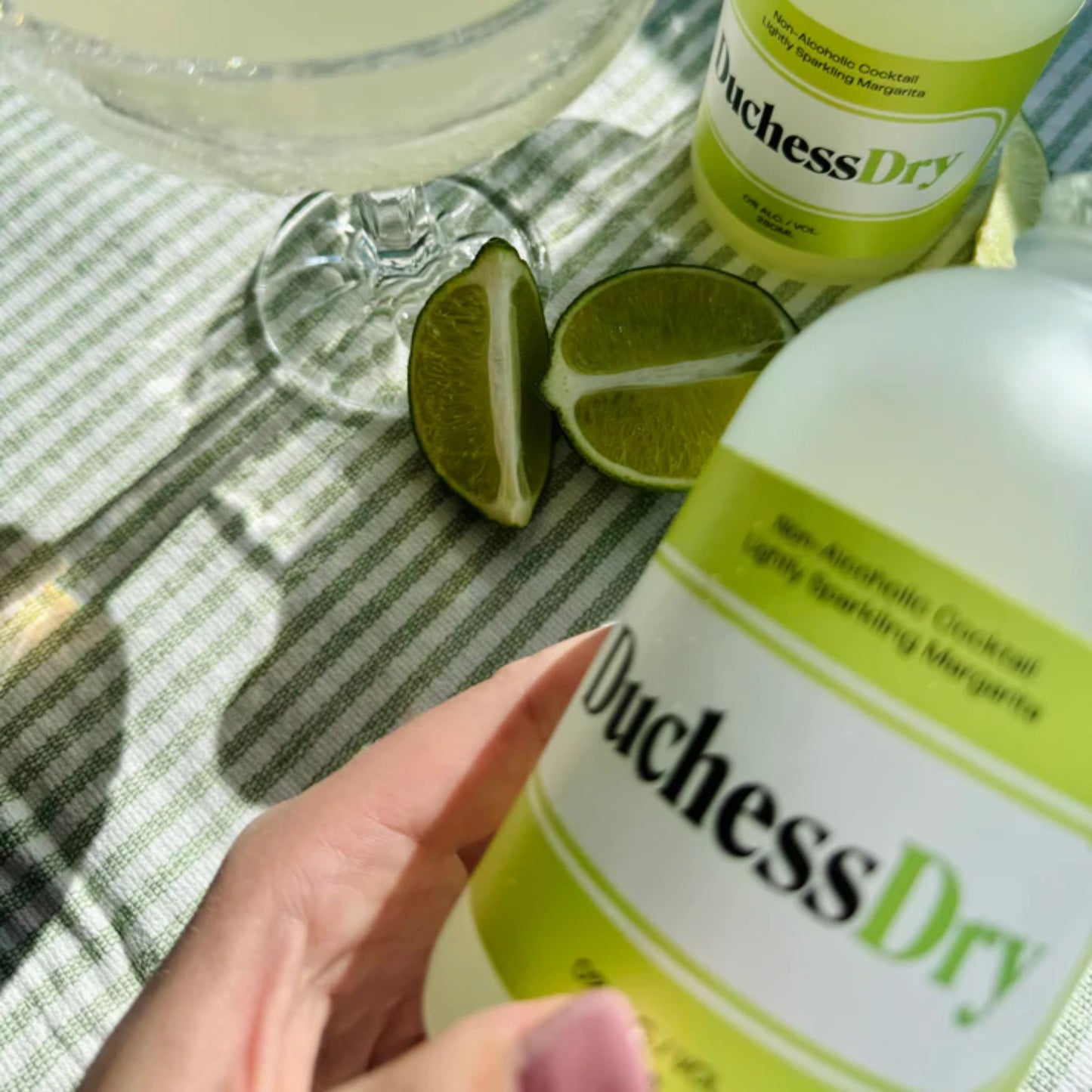 The image shows a hand holding a bottle of DuchessDry Non-Alcoholic Cocktail, specifically the Lightly Sparkling Margarita flavor. Another bottle is on the table resting on a green and white striped tablecloth, with a cocktail glass containing the same drink and lime wedges in the background. The bottles are frosted glass with black and green labels prominently displaying the DuchessDry logo. The overall impression is one of refreshment and a summery vibe.