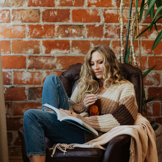 Person relaxing in a cozy chair, reading a book and holding a non‑alcoholic drink against a warm brick wall - KNYOTA Books and Blind Date With a Book vibe.
