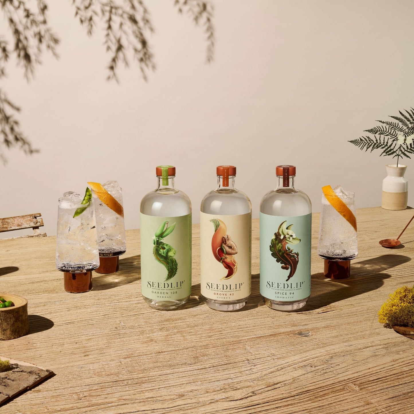 A photo of three 700 milliliter bottles of Seedlip distilled non-alcoholic spirits surrounded by herbs, fruits, and bar tools. Seedlip is available for sale at knyota.com