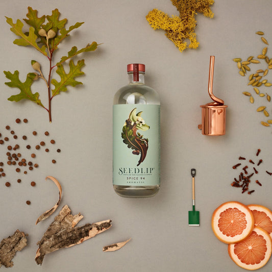 A photo of a 700 milliliter bottle of Seedlip Spice 94 Aromatic surrounded by herbs, fruit, and bar tools. Seedlip is available for sale at knyota.com